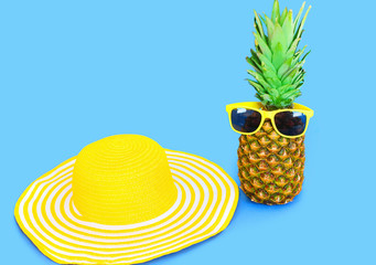 Pineapple with sunglasses and yellow straw beach hat on blue bac