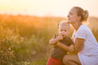© goodmoments - Young beautiful mother hugging her little toddler son on a sunset in a wild flowers field. Happy woman with her baby boy on a summer sunny day. Family walking on the meadow.