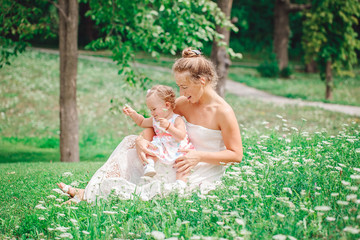 Group of two people, white Caucasian mother and baby girl child in white dress sitting playing in green summer park forest outside, looking in camera,  lifestyle happy childhood concept