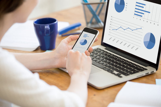 Close-up rear view of young business woman working in office interior on pc holding smartphone and looking at screen with diagrams. Office person using mobile phone and laptop