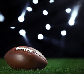 Canvas Print - American football ball on a green grass on dark background and spotlight with laser rays