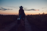 Fototapeta  - Girl in a cowboy hat standing with a suitcase on the road in the sunflower field. Waiting for help. Sunset.