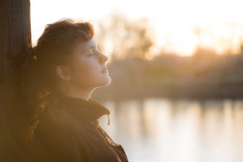 Side Profile Of Peaceful Nirvana Young Woman In Coat With Closed Eyes By Lake With Soft Brown Autumn Light