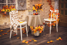 A Cozy Patio. Autumn Leaves Lying On The Wooden Floor, At The Center Are Two White Chairs And A Table With Burlap, Fruits And A Vase Of Berries And Foliage. Background Is Brick Wall And Vintage Window