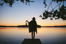 Girl In A Poncho And A Hat With Lamp Standing Back To The View Of The Sunset On The Lake