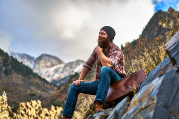 happy bearded guy sitting on luggage at top of mountain