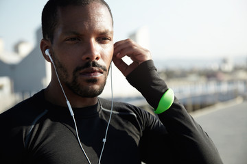 Outdoor shot of attractive muscular male athlete resting after physical activities in open air. Young jogger in black outfit looking into distance, listening to meditative music in earphones