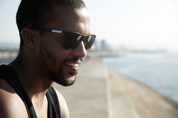 Wall Mural - Charismatic dark-skinned guy in trendy sunglasses standing in morning sun admiring the beginning of day, smiling remembering something funny, against blurred background of big city landscape