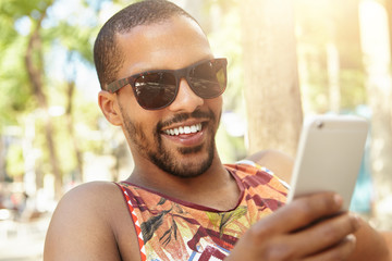 Attractive handsome African radio DJ with sly face expression typing message to his girlfriend with joke or tease using smart phone, smiling while enjoying sunny weather, sitting on bench in park