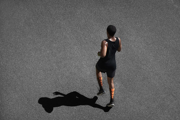 Wall Mural - People, sports and achievement. Top shot of athletic dark-skinned runner wearing black sportswear, jogging outdoors, casting shadow upon black pavement, training, preparing for serious marathon