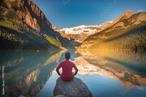 Fotografie, Obraz Man sit on rock watching Lake Louise reflections