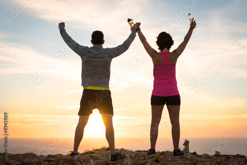 Fotografía Sporty runner couple rising arms in victory sign after successful training outdoor on the coast facing the sun showing back