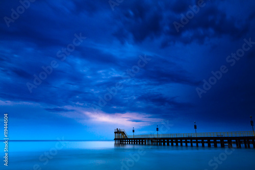 Cuadros en Lienzo Costa de arena con cielo dramático y muelle durante la puesta del sol