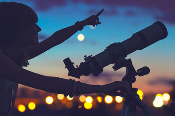 Wall Mural - girl looking at the stars with telescope beside her and de-focused city lights.