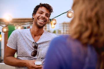 Multi-ethnic millenial couple flirting while having a drink on rooftop terrasse at sunset