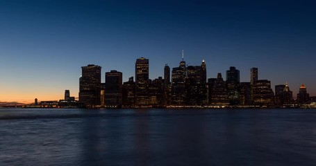 Wall Mural - New York City Lower Manhattan skyscrapers between sunset, dusk and nightfall. Time lapse cityscape view of the Financial District lights and East River with passing boats