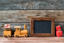 Merry Christmas Decorations, Empty Chalkboard With Toy Wooden Train And Christmas Gifts On A Timber Background