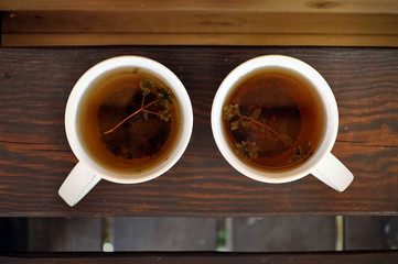 Top view of two white cups with black tea and thyme on a dark wooden surface.