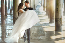 Bride Lifts And Carries Groom In Arms By The Sea Water Ocean Under Pier