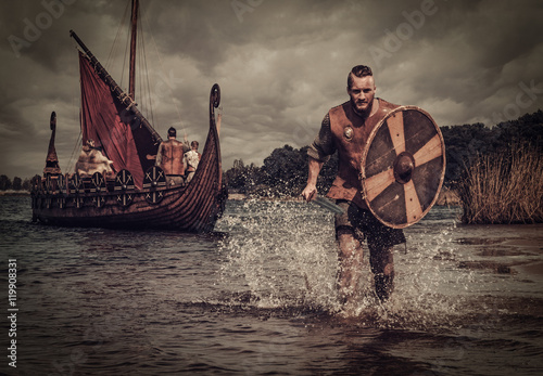 Viking warrior in the attack, running along the shore with Drakkar on the background Canvas Print