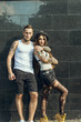 Young stylish tattooed couple standing at the tiled wall on the street looking straight. Boy embracing his girlfriend in the hat with his strong arm. Grunge style concept. Outdoor shot.