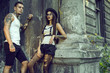 Young stylish tattooed couple in black shorts standing at the column of old ruined building and looking aside. Girl wearing hat, torn tights and glasses. Grunge style concept. Outdoor shot. Copy-space