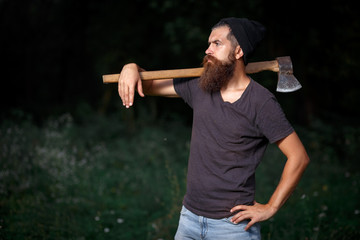 Wall Mural - Brutal brunette bearded man in warm hat with a hatchet in the woods on a background of trees