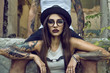 Gorgeous tattooed girl with provocative make up sitting between her boyfriend's legs in the ruined abandoned building, her arms on his knees. She is wearing black hat and glasses. Grunge style concept