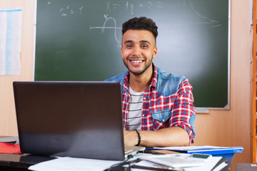 Hispanic Man Using Laptop Computer, Student In University Classroom At Desk Over Chalk Board