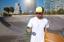 Closeup on surfer in trucker cap and sunglasses, walking with his longboard in hand and looking at his smartphone while listening music through earplugs headphones in urban skatepark, front view
