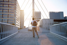 A Traveler In Khakis, White T-shirt, Fedora Hat Walks Away On A City Bridge At Sunset