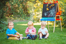 Group Of Three White Caucasian Toddler Children Kids Boys And Girl Sitting Outside In Summer Autumn Park By Drawing Easel With Books, Reading Studying Learning, Back To School