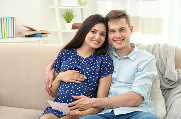 Poster - Beautiful young pregnant couple with ultrasound image sitting on sofa