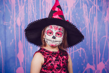 Little Girl Dressed As  Witch With Makeup On Her Face Standing   Background Of Purple Wall