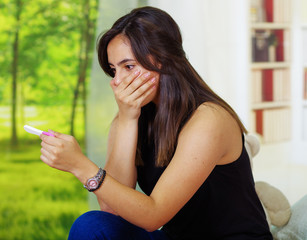 Wall Mural - Pretty young brunette woman holding pregnancy home test, shocked facial expression covering mouth with hand, bookshelves interior garden window background