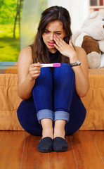 Wall Mural - Pretty young brunette woman sitting on floor holding pregnancy home test, crying wiping tears, looking emotional, garden window background