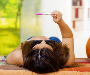 Wall Mural - Pretty young brunette woman lying down holding up pregnancy home test in front, as seen from behind head, bookshelves and window garden background