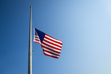 American flag flies at half mast backlit by the sun in bright blue sky