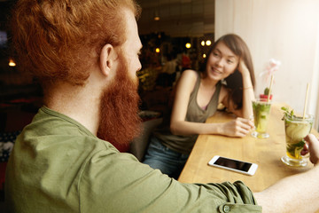 Wall Mural - Two friends of different ethnicities talking to each other sitting at coffee shop, having fresh cocktails. Cute Asian woman looking at her hipster companion with curious expression. Selective focus