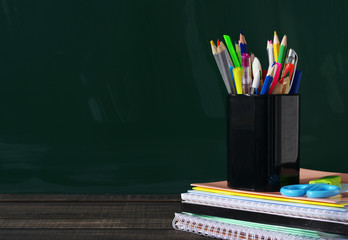 Wall Mural - School supplies on a wooden surface against a blackboard. Books, notebooks, handles, colored pencils and rulers in a glass on a wooden table