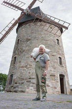 Senior Miller With Flour Sack Posing In Front Of Medieval Windmill