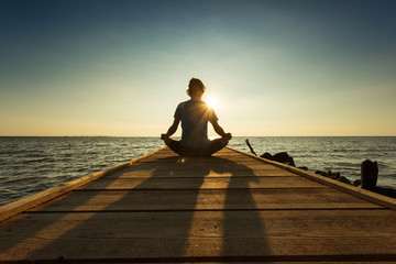 Young man sitting and meditating on a pontoon by a lake at the s