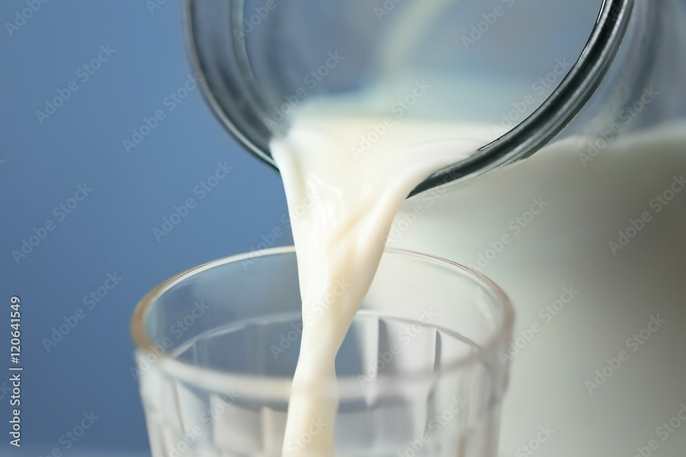 Milk pouring from a jug into a glass on blue background, closeup