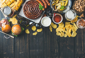 Wall Mural - Beer and snack set. Octoberfest food frame concept. Variety of beers, grilled sausages, burgers, corn, fried potatoes, chips, salted almonds and sauces on dark wooden background. Top view, copy space