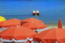 Orange Sun Umbrellas On A Sunny Beach With A Small Boat On The Blue Sea In The Distance