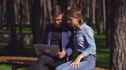 Poster - Man and woman using laptop outdoors. Happy couple on the meeting or date looking on the computer screen smiling. Handsome guy and attractive girl sitting on the bench in the park.