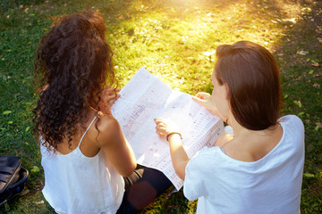 A caucasian woman is pointing at the place on the map while sitting in a city park with a friend. Two young girls are exploring a paper map of a city while sitting on a lawn. View from the top.