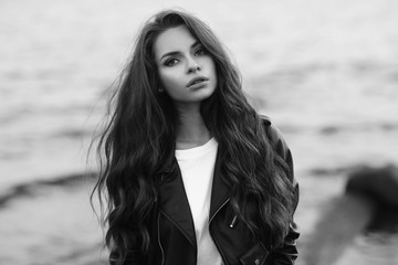 Young pretty stylish girl in black leather jacket posing, walking or standing at beach against water in bokeh. Fashionable woman with long curly hair looking at you.