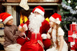 © pressmaster - An image of Santa Claus sitting at fireplace and Christmas tree and giving presents to two children
