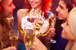 © pressmaster - A close up of four clinking glasses against the girl holding her birthday cake among her friends